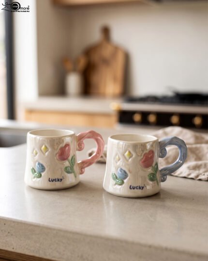 Two glazed ceramic mugs with embossed 3D flowers, stars, and pastel pink and blue artistic handles placed on a kitchen counter.