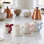 Four glossy ceramic coffee mugs with decorative bow handles in red, white, pink, and blue, arranged on a marble kitchen counter.