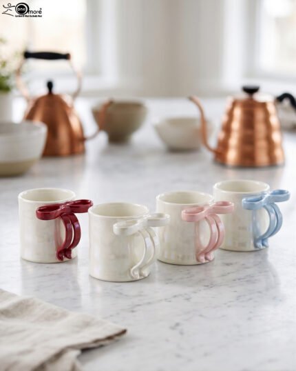 Four glossy ceramic coffee mugs with decorative bow handles in red, white, pink, and blue, arranged on a marble kitchen counter.