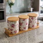 Three floral print ceramic storage jars with bamboo lids arranged on a matching wooden tray on a modern kitchen countertop