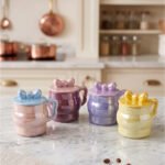 Four pastel ceramic mugs with matching lids and cute bow details, featuring a glossy pearl finish arranged on a marble kitchen countertop.