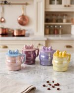 Four pastel ceramic mugs with matching lids and cute bow details, featuring a glossy pearl finish arranged on a marble kitchen countertop.