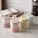 Four pastel ceramic mugs with animal embossed patterns, matching lids with round knobs, and spoons displayed on a kitchen counter