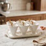 White ceramic ice cream set with 6 swirled pedestal bowls, matching serving tray, and gold leaf spoons displayed on a marble kitchen counter.