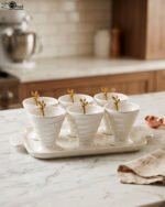 White ceramic ice cream set with 6 swirled pedestal bowls, matching serving tray, and gold leaf spoons displayed on a marble kitchen counter.