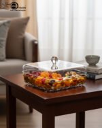 Two square crystal dry fruit dishes with gold trim bases and glass lids displayed on a wooden table in a luxury living room setting.