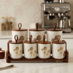 White ceramic kitchen storage jars with gold dandelion print, wooden lids with loop handles, and a wooden rack on a kitchen counter.
