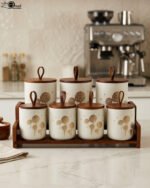 White ceramic kitchen storage jars with gold dandelion print, wooden lids with loop handles, and a wooden rack on a kitchen counter.