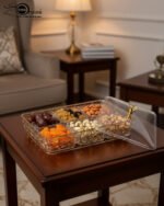 Clear acrylic dry fruit serving tray with gold-toned wire compartments, a matching lid, and filled with assorted nuts and dried fruits on a wooden table.