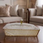 Elegant partitioned glass dry fruit dish with a gold trim and crystal-accented lid sitting on a wooden coffee table in a modern living room.