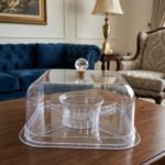 Clear acrylic compartmentalized dry fruit and cake serving dish with a square transparent lid and crystal handle on a wooden table.