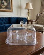 Clear acrylic compartmentalized dry fruit and cake serving dish with a square transparent lid and crystal handle on a wooden table.