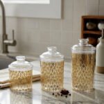 Three clear acrylic storage jars of varying heights featuring a decorative gold pattern and matching lids, displayed on a kitchen countertop next to spices.