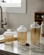 Three clear acrylic storage jars of varying heights featuring a decorative gold pattern and matching lids, displayed on a kitchen countertop next to spices.