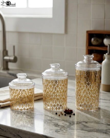Three clear acrylic storage jars of varying heights featuring a decorative gold pattern and matching lids, displayed on a kitchen countertop next to spices.