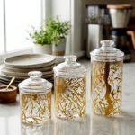 Three transparent acrylic storage jars of varying heights featuring a decorative gold branch pattern, arranged on a marble kitchen countertop with plates and a plant in the background.