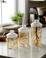 Three transparent acrylic storage jars of varying heights featuring a decorative gold branch pattern, arranged on a marble kitchen countertop with plates and a plant in the background.