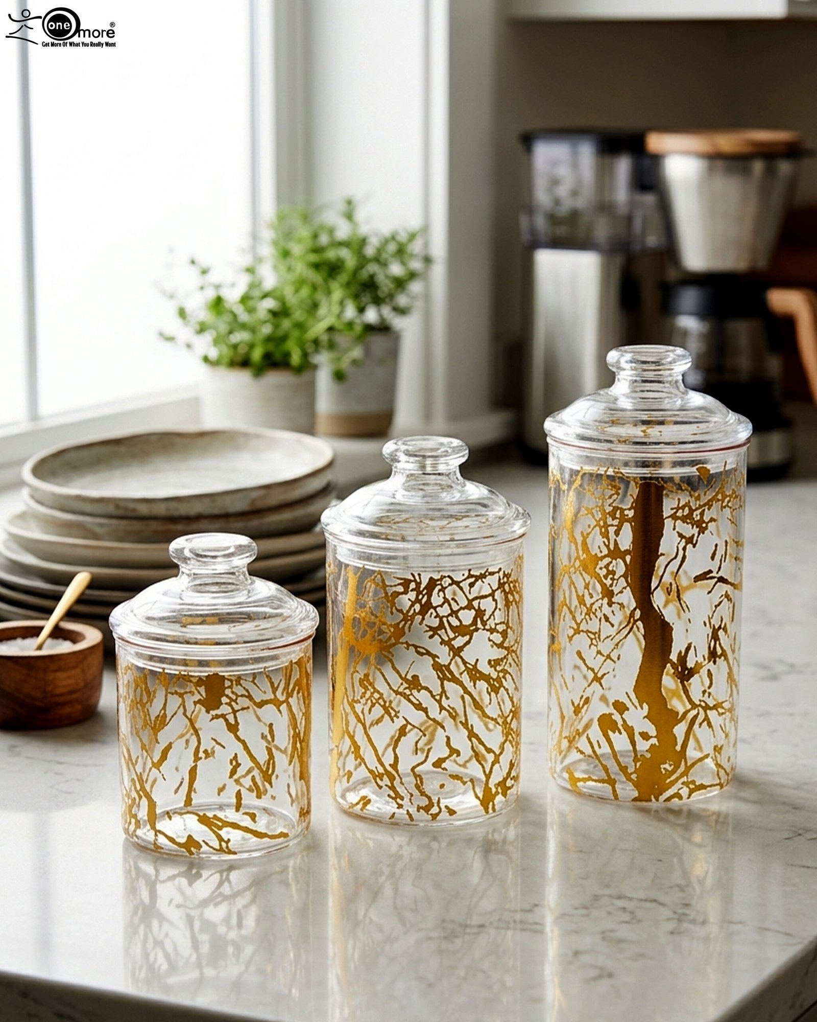 1 Three transparent acrylic storage jars of varying heights featuring a decorative gold branch pattern, arranged on a marble kitchen countertop with plates and a plant in the background.