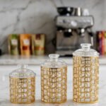 Three luxurious clear acrylic jars of varying heights featuring intricate gold geometric designs and matching lids, displayed neatly on a white marble countertop with a small golden spoon.