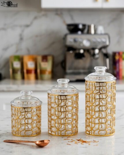 Three luxurious clear acrylic jars of varying heights featuring intricate gold geometric designs and matching lids, displayed neatly on a white marble countertop with a small golden spoon.