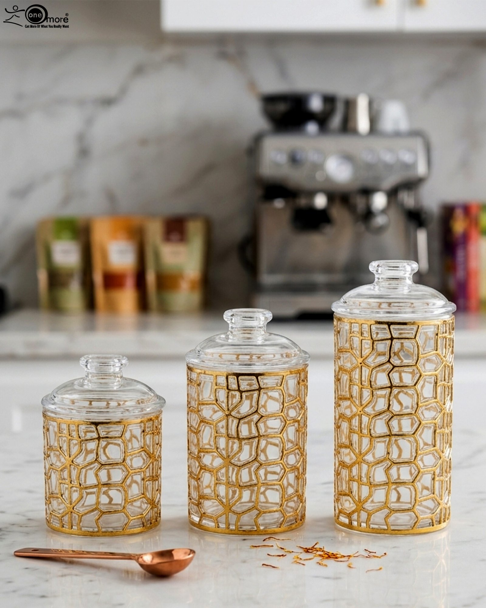 1 Three luxurious clear acrylic jars of varying heights featuring intricate gold geometric designs and matching lids, displayed neatly on a white marble countertop with a small golden spoon.