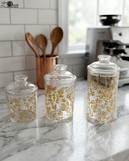 Three clear acrylic jars of varying heights featuring a gold floral print and clear lids, arranged on a marble kitchen countertop.