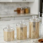 Three luxury clear acrylic jars with gold horizontal stripe patterns and matching lids displayed on a marble countertop in a modern kitchen.