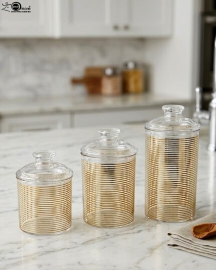 Three luxury clear acrylic jars with gold horizontal stripe patterns and matching lids displayed on a marble countertop in a modern kitchen.