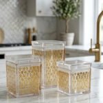 Set of three clear acrylic kitchen storage jars with elegant gold patterned designs, displayed on a marble countertop.