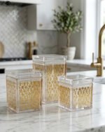 Set of three clear acrylic kitchen storage jars with elegant gold patterned designs, displayed on a marble countertop.