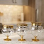 Three graduated luxury clear acrylic storage jars on pedestals with gold bases, matching gold lids, silver locking latches, and spoons arranged on a marble countertop.