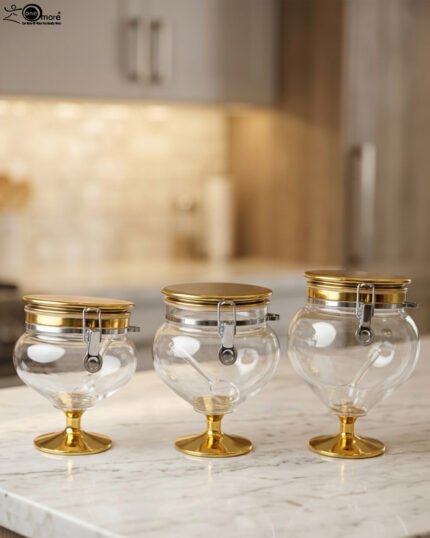 Three graduated luxury clear acrylic storage jars on pedestals with gold bases, matching gold lids, silver locking latches, and spoons arranged on a marble countertop.