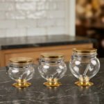 Three graduated clear acrylic jars with gold bases, lids, and matching spoons displayed on a dark countertop from One More Crockery.
