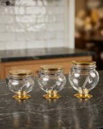 Three graduated clear acrylic jars with gold bases, lids, and matching spoons displayed on a dark countertop from One More Crockery.
