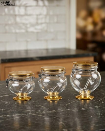 Three graduated clear acrylic jars with gold bases, lids, and matching spoons displayed on a dark countertop from One More Crockery.