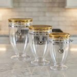 Three clear luxury acrylic storage jars of varying sizes featuring gold lids and secure silver airtight locking clasps, arranged on a marble countertop in a modern kitchen.