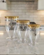Three clear luxury acrylic storage jars of varying sizes featuring gold lids and secure silver airtight locking clasps, arranged on a marble countertop in a modern kitchen.