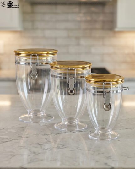 Three clear luxury acrylic storage jars of varying sizes featuring gold lids and secure silver airtight locking clasps, arranged on a marble countertop in a modern kitchen.