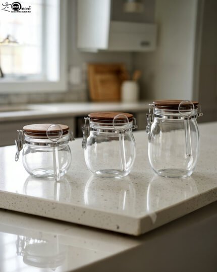 Three transparent acrylic airtight storage jars of varying sizes featuring wooden lids, metal clamp locks, and attached spoons, displayed neatly on a white kitchen countertop from One More Crockery.