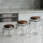 Three clear acrylic storage jars of varying sizes featuring wooden lids, airtight wire-clamp seals, and small hanging spoons, arranged on a white marble countertop in a modern kitchen.