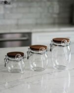 Three clear acrylic storage jars of varying sizes featuring wooden lids, airtight wire-clamp seals, and small hanging spoons, arranged on a white marble countertop in a modern kitchen.