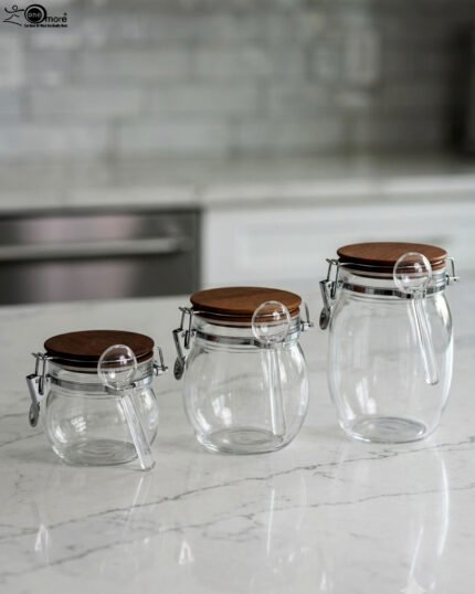 Three clear acrylic storage jars of varying sizes featuring wooden lids, airtight wire-clamp seals, and small hanging spoons, arranged on a white marble countertop in a modern kitchen.