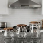 Clear acrylic kitchen storage jar set of three in varying sizes with wooden lids, airtight clasp locks, and attached spoons sitting on a dark marble countertop against a modern kitchen background.