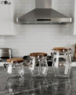 Clear acrylic kitchen storage jar set of three in varying sizes with wooden lids, airtight clasp locks, and attached spoons sitting on a dark marble countertop against a modern kitchen background.