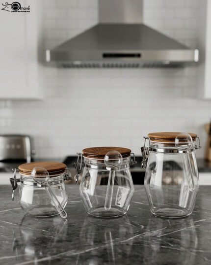 Clear acrylic kitchen storage jar set of three in varying sizes with wooden lids, airtight clasp locks, and attached spoons sitting on a dark marble countertop against a modern kitchen background.