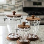Set of three clear acrylic jars of varying sizes with wooden bases, wooden lids, airtight metal clamps, and hanging acrylic spoons on a kitchen countertop.