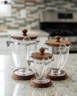 Set of three clear acrylic jars of varying sizes with wooden bases, wooden lids, airtight metal clamps, and hanging acrylic spoons on a kitchen countertop.