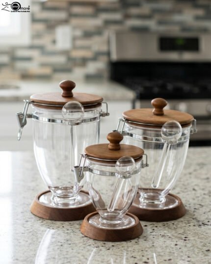 Set of three clear acrylic jars of varying sizes with wooden bases, wooden lids, airtight metal clamps, and hanging acrylic spoons on a kitchen countertop.