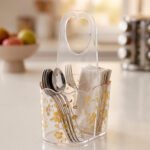 Clear acrylic spoon and cutlery holder featuring a gold floral design, divided compartments holding silverware and napkins, and a heart-shaped handle on a kitchen countertop