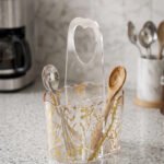 Clear acrylic spoon holder and cutlery caddy with gold accent patterns and a heart-shaped handle on a kitchen countertop holding utensils.